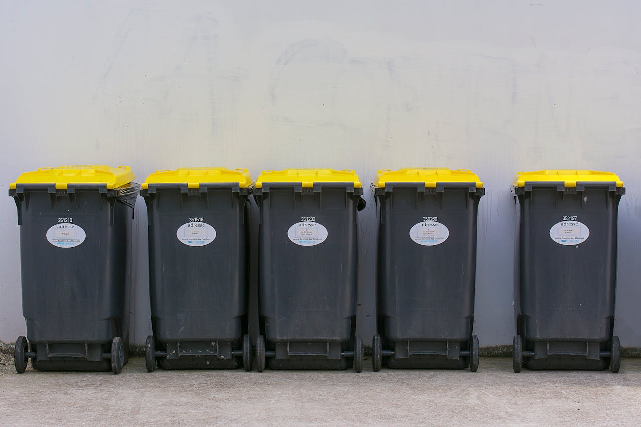 Neatly lined up garbage bins with yellow lids against a plain background wall.