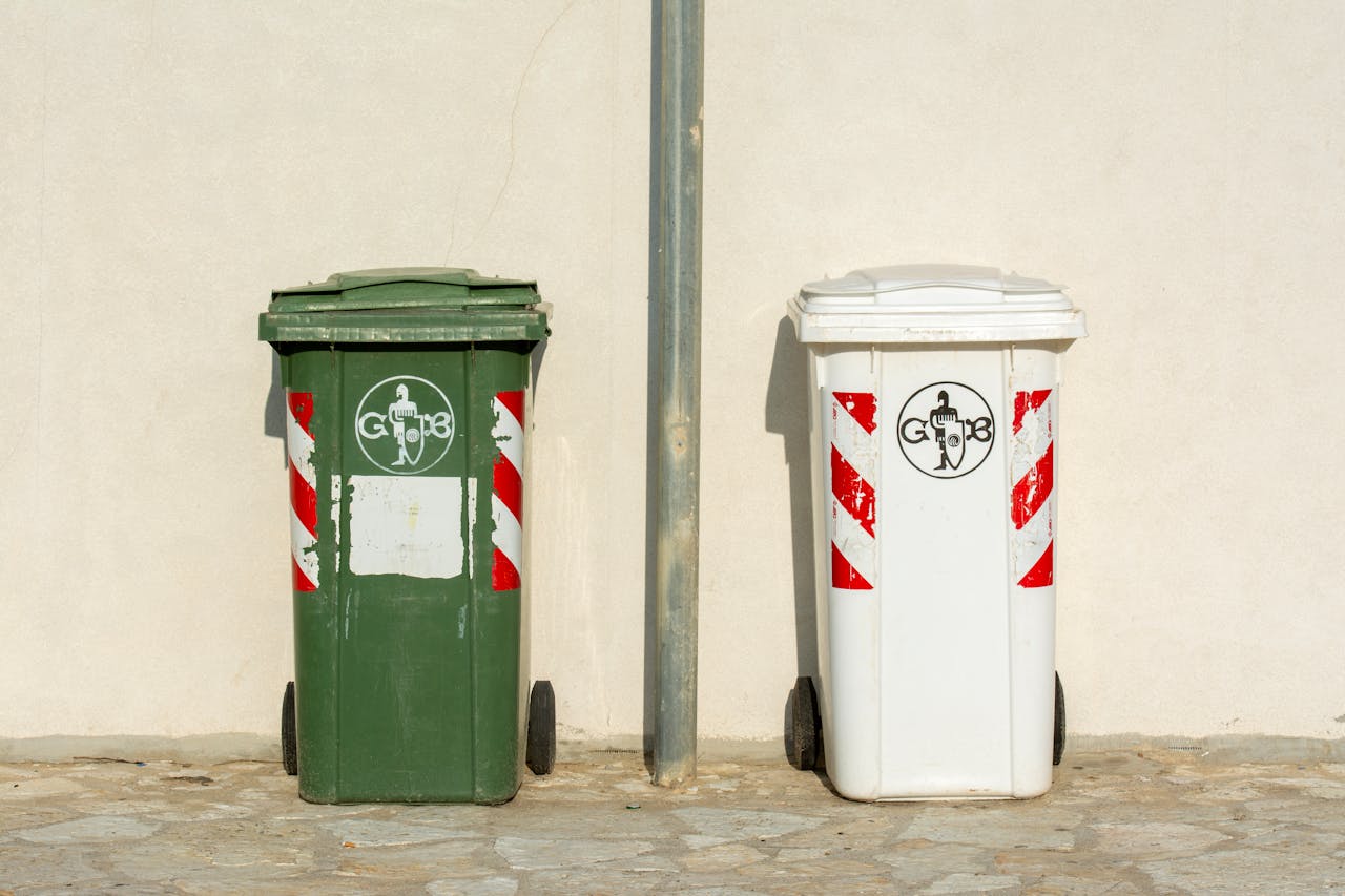 Two outdoor garbage bins on a sidewalk, one green and one white, against a wall.